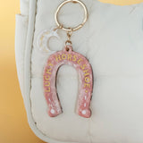 Pink horseshoe keychain with 'Lucky Horseshoe' text on a white bag against a yellow background