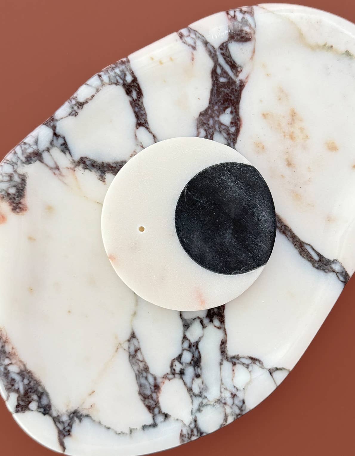A close-up of a white circular stone incense holder with a black circular inset, displayed on a white and brown marbled surface against a warm terracotta background.