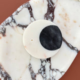 A close-up of a white circular stone incense holder with a black circular inset, displayed on a white and brown marbled surface against a warm terracotta background.