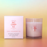 A SC ORP IO scented candle in a frosted glass jar next to its white packaging box, featuring pink and orange text, set against a pastel gradient background.