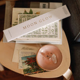 Stack of books with a 'Moon Glow' bookmark on a small table next to a pink candle.