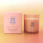 A pink scented candle in a frosted clear glass vessel stands beside its matching pink box on a soft gradient background. The packaging and glass both show the brand name Sag Ita Rius and scent text including Cardamom, Pear, and Sandalwood.
