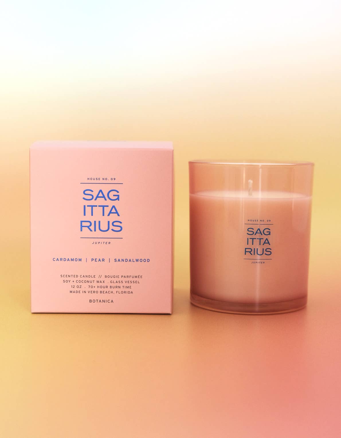 A pink scented candle in a frosted clear glass vessel stands beside its matching pink box on a soft gradient background. The packaging and glass both show the brand name Sag Ita Rius and scent text including Cardamom, Pear, and Sandalwood.