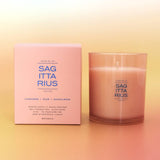 A pink scented candle in a frosted clear glass vessel stands beside its matching pink box on a soft gradient background. The packaging and glass both show the brand name Sag Ita Rius and scent text including Cardamom, Pear, and Sandalwood.