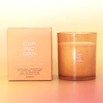 A scented candle in a frosted amber glass jar sits next to its matching peach-colored packaging box. The brand name 'CAP RIC ORN' is clearly printed on both the box and the jar.
