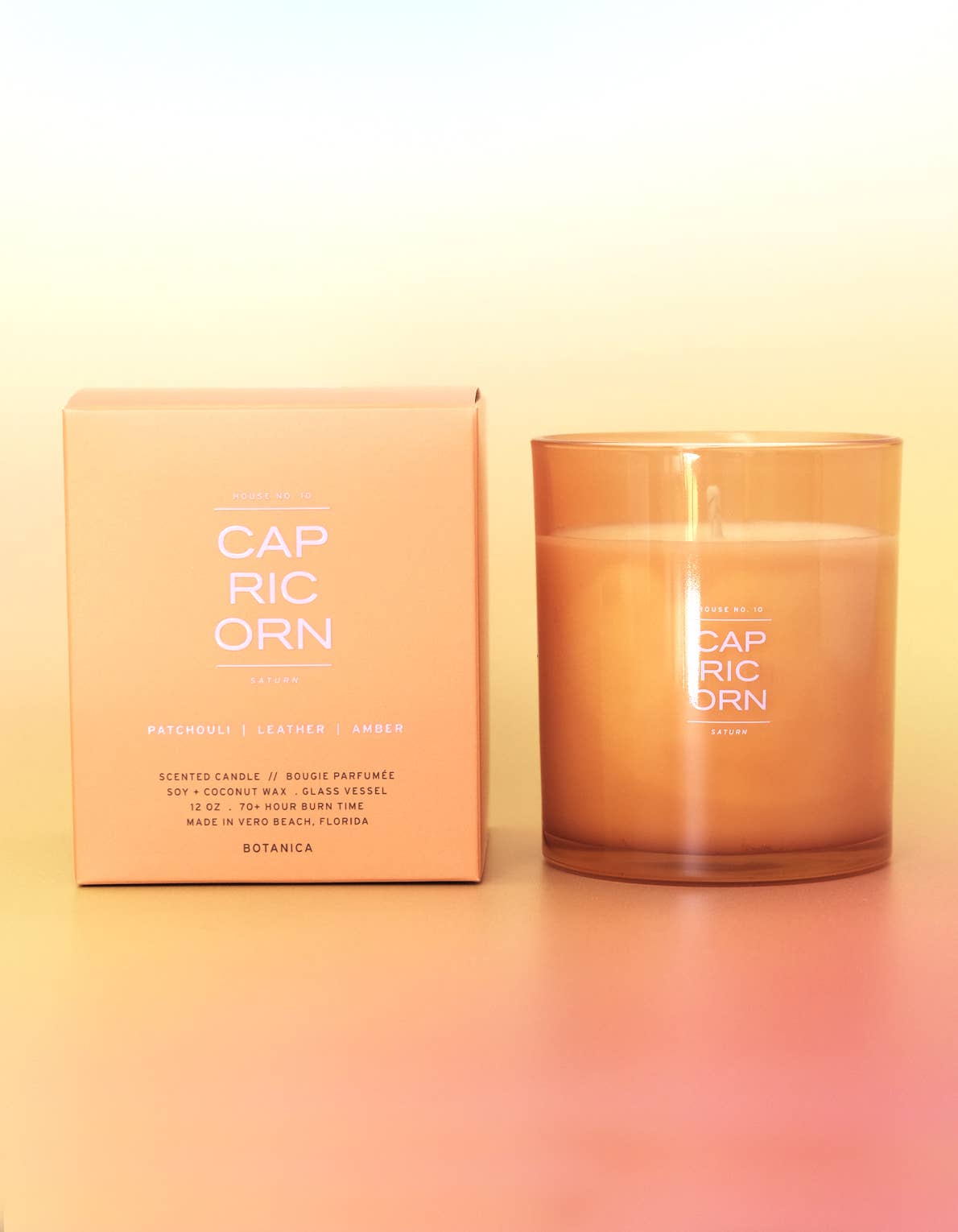 A scented candle in a frosted amber glass jar sits next to its matching peach-colored packaging box. The brand name 'CAP RIC ORN' is clearly printed on both the box and the jar.