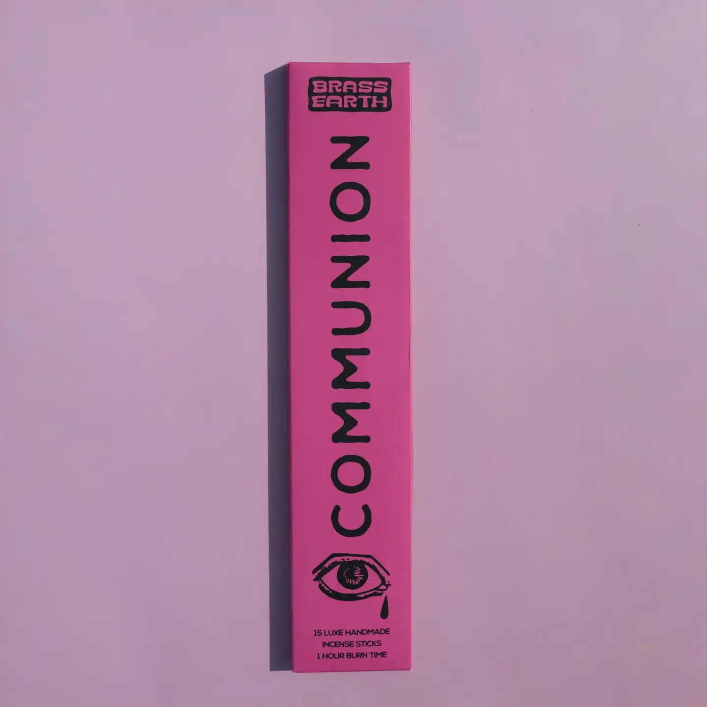 A tall, narrow pink box for Brass Earth Communion incense sticks, featuring black text and an eye graphic, set against a solid lavender-pink background.