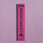 A tall, narrow pink box for Brass Earth Communion incense sticks, featuring black text and an eye graphic, set against a solid lavender-pink background.