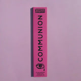 A tall, narrow pink box for Brass Earth Communion incense sticks, featuring black text and an eye graphic, set against a solid lavender-pink background.