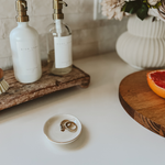 Jewelry dish with rings on a white surface, with bottles and a fruit in the background.
