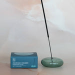 Green incense holder with a single stick of incense next to its packaging on a light background