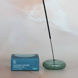 Green incense holder with a single stick of incense next to its packaging on a light background