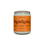 A glass jar candle with a bright orange label reading 'Samhain' and 'Ghast Poppy' on a soft pink studio background.