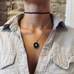 Person wearing a gold necklace with a black evil eye pendant against a stone wall background