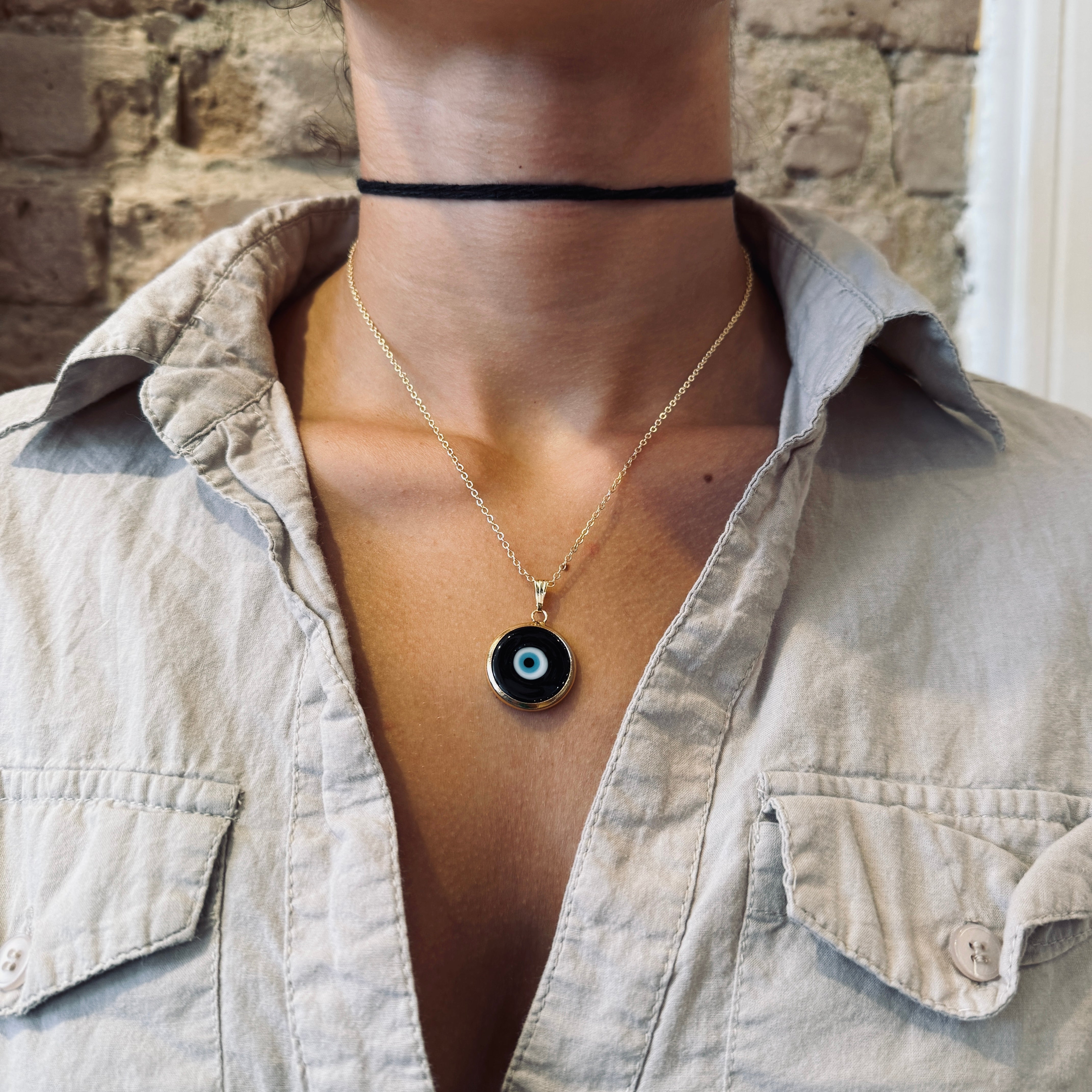 Person wearing a gold necklace with a black evil eye pendant against a stone wall background